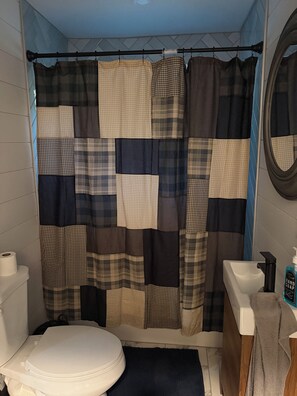 A bathroom with a patchwork shower curtain, a toilet, a sink, and a mirror. - Happy Acre of Wolfpen a charming little 2 bed 1 bath home (Mena)