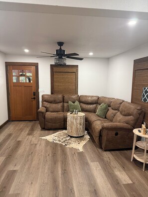 Ein Wohnzimmer mit einem braunen Ledersofa, einem runden Holztisch, einem Deckenventilator und einer Holztür.