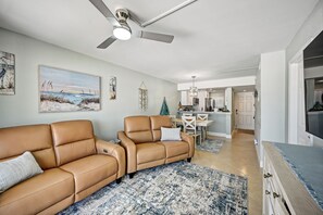 Ein Wohnzimmer mit beiger Couch, einem Essbereich mit Tisch und Stühlen, einer Küche im Hintergrund und einem Deckenventilator.