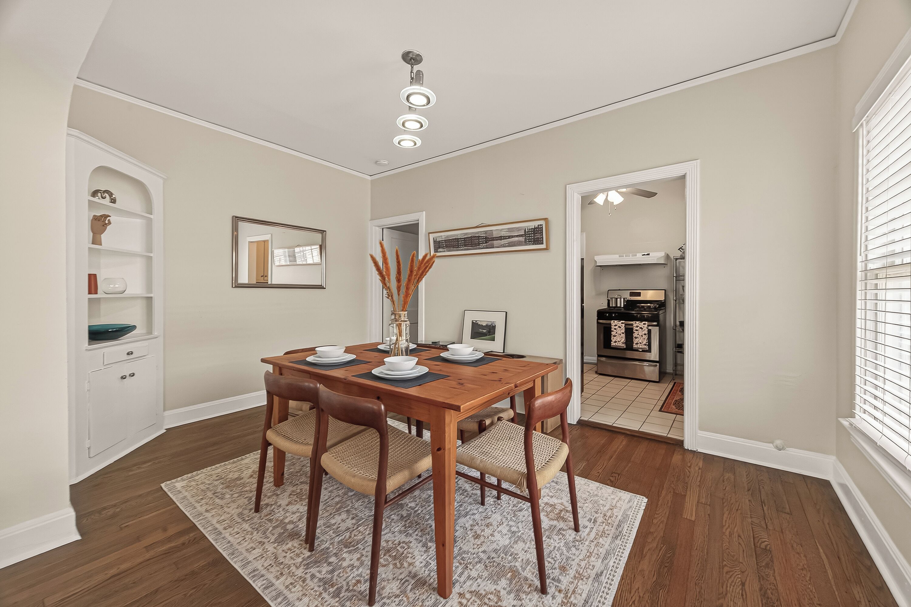 A dining area with a wooden table, four chairs, a rug, a ceiling light fixture, a white cabinet, and a framed picture on the wall.