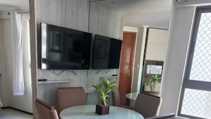 Een eetgedeelte met een ronde tafel, zes stoelen, een potplant en een flatscreen televisie aan de muur.
