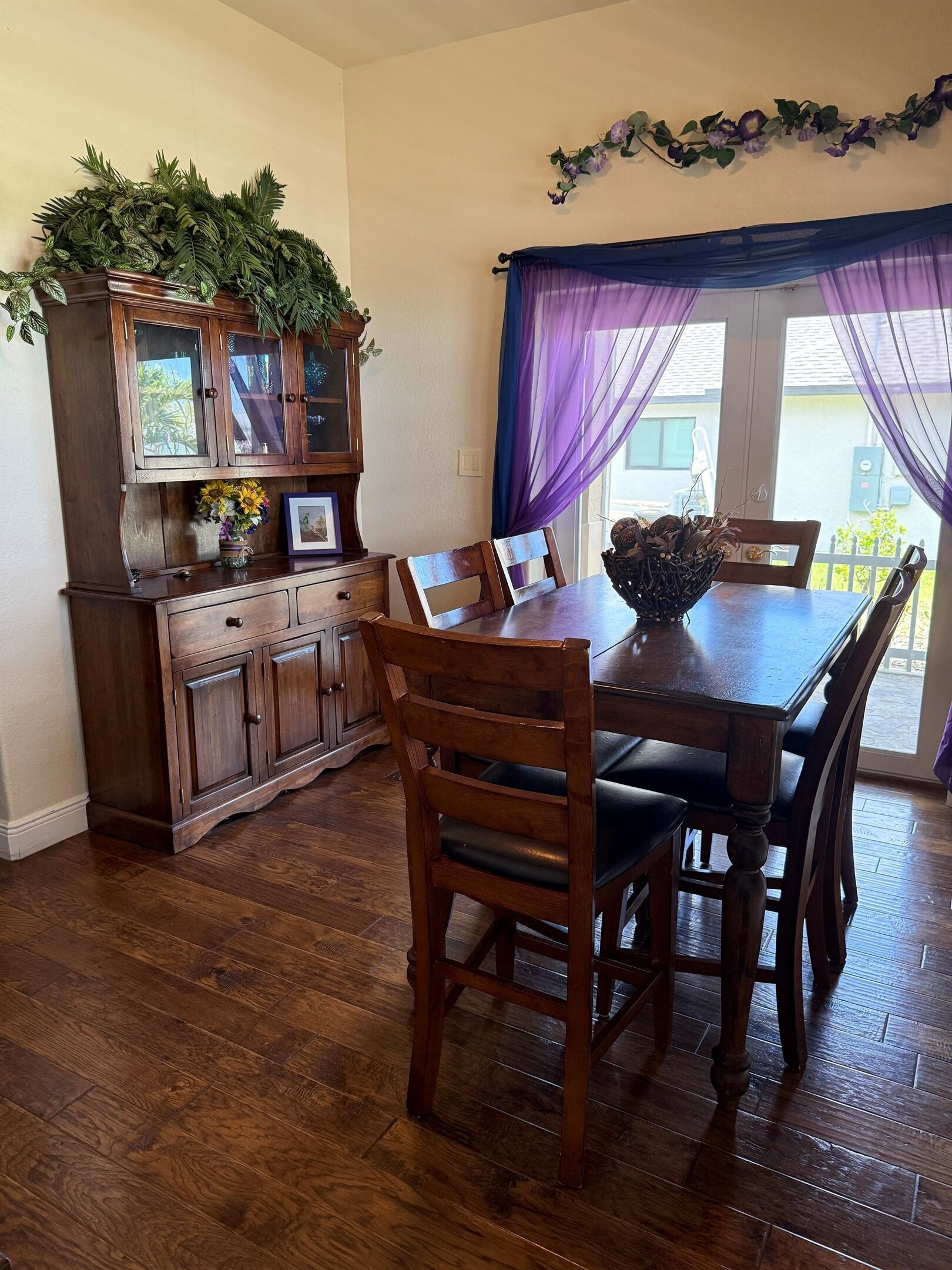 A dining area with a wooden table and chairs, a cabinet with glass doors, and a window with purple curtains.