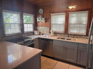 A kitchen with wooden cabinets, a stove, a sink, and a window with blinds. - Sand in our Shoes | Coastal comfort just steps from the shore 🌊 (North Myrtle Beach)