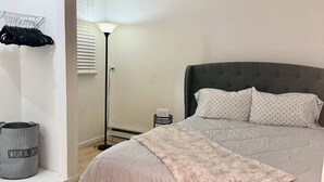 Ein Schlafzimmer mit einem Bett, einem Nachttisch, einer Lampe, einem Wandregal mit Kleidung und einer Mülltonne.