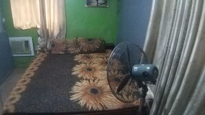 Una habitación con cama, ventilador de pie, ventana con cortinas y aire acondicionado instalado en la pared.