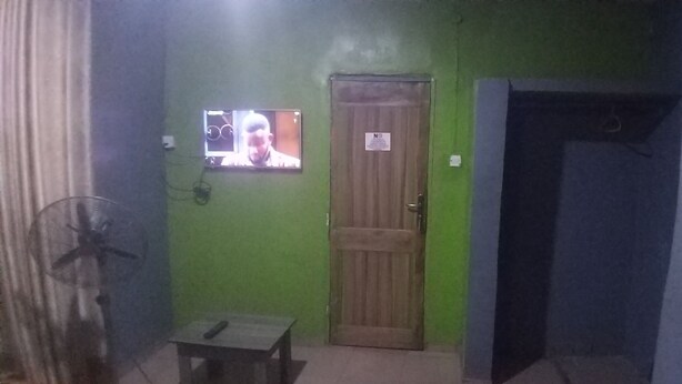 Una habitación con pared verde, puerta de madera, una mesita, un ventilador y una pantalla de televisión que muestra a una persona.