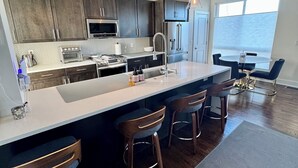 Une cuisine moderne avec des meubles de couleur sombre, des appareils en acier inoxydable, un îlot central doté de tabourets, un éclairage suspendu et une grande fenêtre.