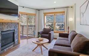 A living room with a stone fireplace, a brown sofa, a round coffee table, and large windows offering a mountain view. - New Listing! Stylish new design, ski in/out. Watch World Cup, view golf course! (Frisco)