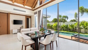 Sala de estar moderna com uma televisão grande, área de jantar com mesa de vidro e vista para uma piscina e vegetação.