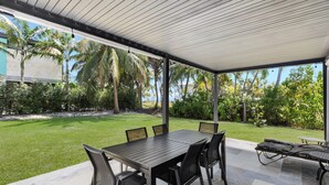 An outdoor patio with a table and chairs, a covered area, and a view of a well-maintained lawn and palm trees. - Surf Shack - Downtown Islamorada 2 Bedroom Beach Bungalow (Islamorada)