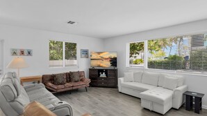 A modern living room with a grey sofa, a brown leather sofa, and a white sofa. There is a TV on a wooden cabinet, and the room has large windows with blinds. - Surf Shack - Downtown Islamorada 2 Bedroom Beach Bungalow (Islamorada)
