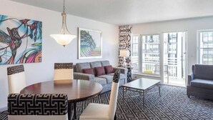 Ein modernes Wohnzimmer mit einer Couch, einem Esstisch mit gemusterten Stühlen, einer Pendelleuchte und großen Fenstern mit Blick auf Gebäude.