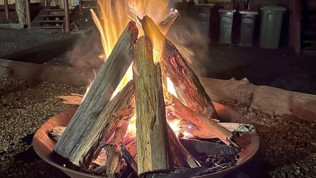 A fire pit with logs, surrounded by gravel, with a building and a staircase in the background. - Froggy’s Quarters Cabin 5 - Koala (Halls Gap)
