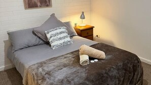 A bedroom with a bed, pillows, a bedside lamp, a window, and a wall-mounted picture. - Froggy’s Quarters Cabin 5 - Koala (Halls Gap)