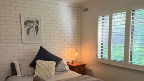 Un dormitorio con pared de ladrillo, una cama con muchos cojines, una mesita de noche con lámpara y una ventana con persianas.