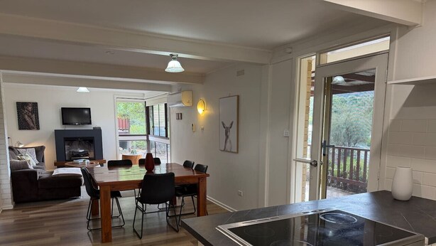 A modern kitchen with a central island, a dining area with a wooden table and chairs, and a living room with a sofa and a television. - Froggy`s Quarters Cabin 1 - Cockatoo (Halls Gap)