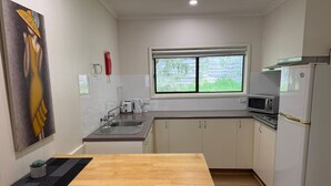 A compact kitchen with a wooden table, white cabinets, a microwave, and a window with a view of greenery. - Froggy’s Quarters Cabin 3 - Emu (Halls Gap)