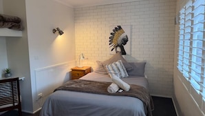 A bedroom with a white brick wall, a bed with grey bedding, a side table with a lamp, and a window with blinds. - Froggy’s Quarters Cabin 3 - Emu (Halls Gap)