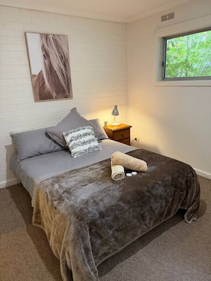 Un dormitorio con cama, almohadas, una lámpara de noche, una ventana y un cuadro en la pared.