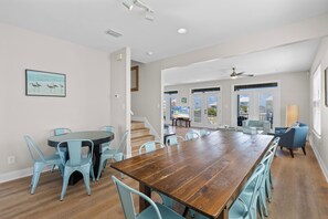 A spacious dining area with a long wooden table and light blue chairs, a staircase leading to a balcony, and a view of the outdoors. - Dog Friendly Beach House 200ft from the Beach (Gulf Shores)