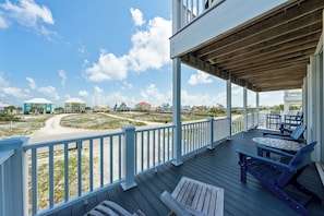 Eine überdachte Terrasse mit Sitzgelegenheiten im Freien, Blick auf den Strand und Gebäude im Hintergrund.