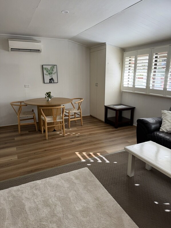 A living room with a wooden dining table, two chairs, a black leather sofa, a small side table, and a window with blinds.