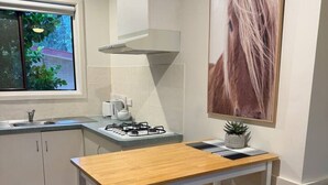 A compact kitchen with a wooden table, a gas stove, a window with a view of greenery, and a large framed picture on the wall. - Froggy’s Quarters Cabin 8 - Possum (Halls Gap)