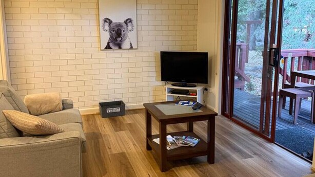 A living room with a sofa, a coffee table, a television, and a large window with a view of outdoor furniture. - Froggy’s Quarters Cabin 8 - Possum (Halls Gap)