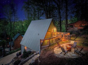 Eine Hütte mit geneigtem Dach, einer Terrasse mit Stühlen und einer Feuerstelle, umgeben von Bäumen.