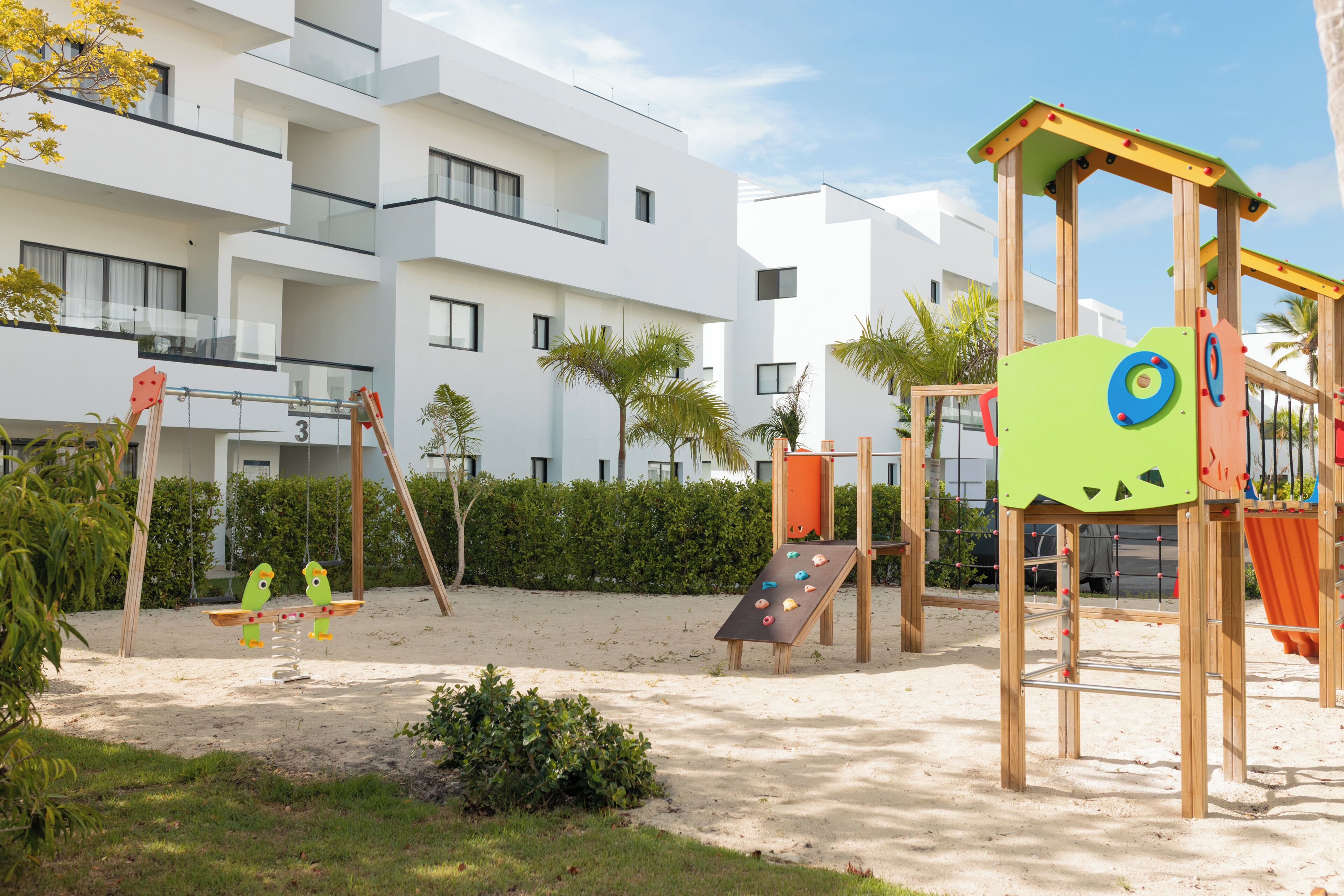 Un parque infantil con columpios y toboganes, rodeado de arena y vegetación.