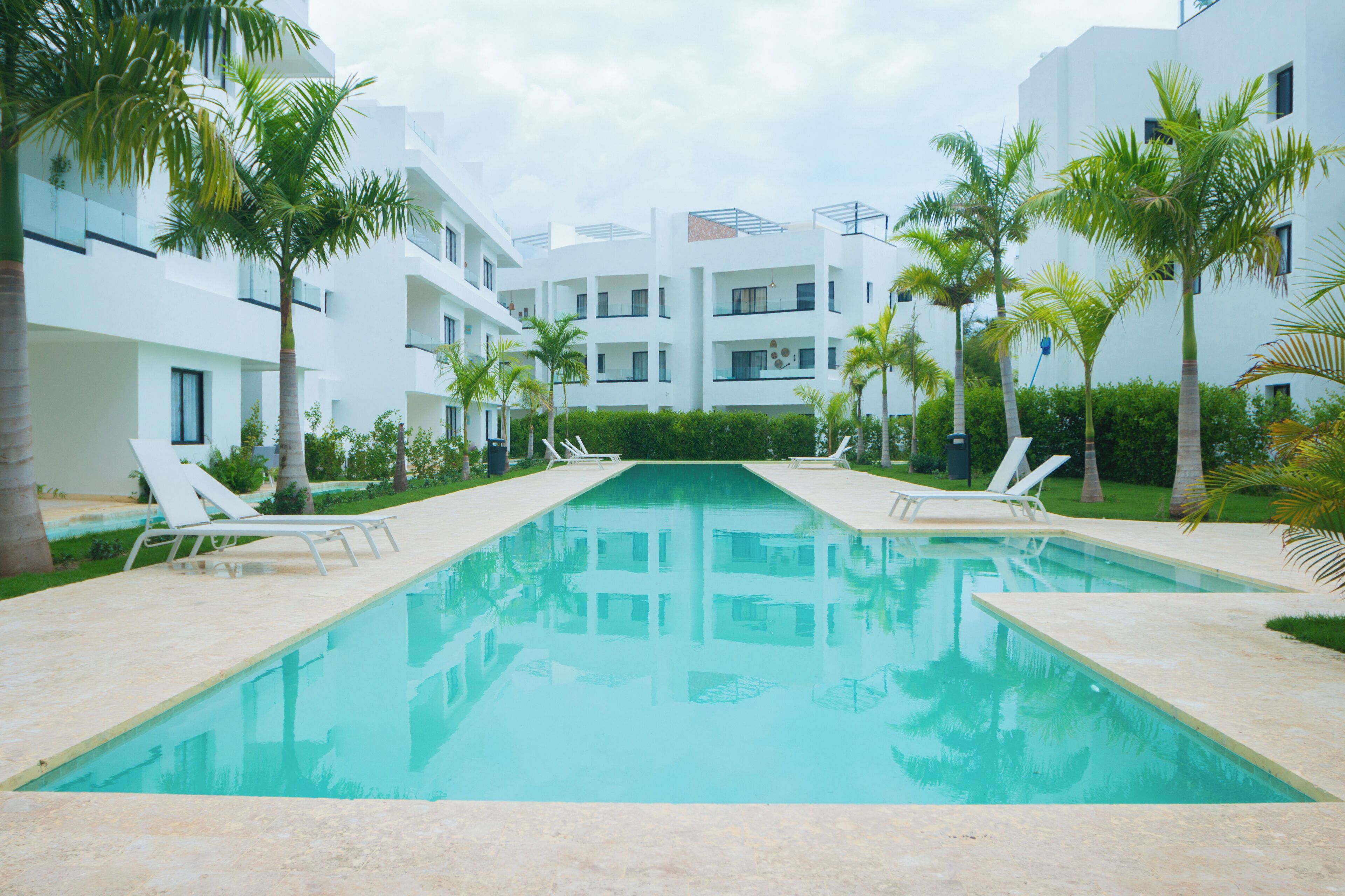 A swimming pool with lounge chairs, surrounded by palm trees and modern buildings.