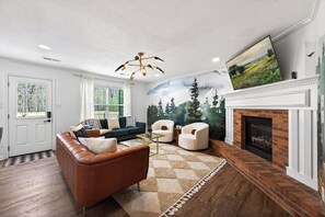 Ein Wohnzimmer mit Kamin, einer Couch, Sesseln, einem Couchtisch und einem Wandgemälde von einem Wald.