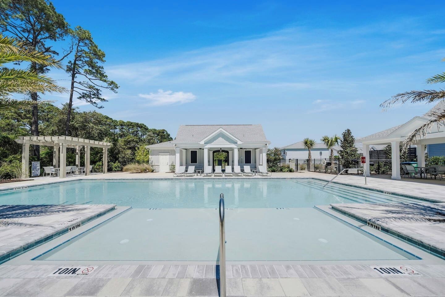 A rectangular pool with a pergola and lounge chairs on one side, a covered patio area with tables and chairs on the other, and a building with a gabled roof in the background.
