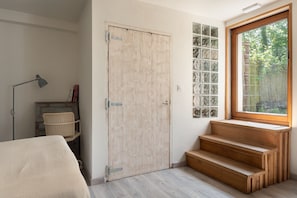 Una habitación con puerta de madera, ventana con vista a follaje, lámpara y una silla.