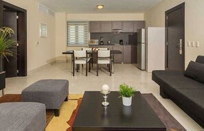Ein modernes Wohnzimmer mit Sofa, Hocker und Couchtisch. Im Hintergrund ist eine Küche erkennbar und links eine Tür.