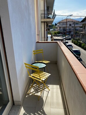 Un balcón con sillas amarillas y una mesa, con vista a una calle con coches estacionados y edificios.