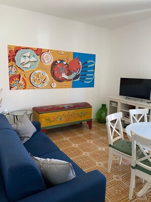 Una sala de estar con un sofá azul, un cuadro de pared colorido, un televisor, una mesita con sillas y un piso estampado.