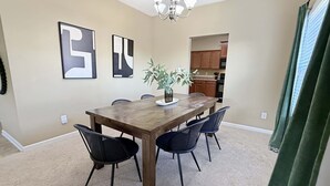 Un comedor con una mesa de madera, cuatro sillas, dos cuadros abstractos, una araña y vista a la cocina.
