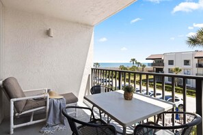 En balkon med et bord og stole, udsigt til havet og moderne bygninger i baggrunden.