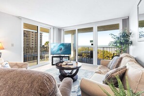 Een woonkamer met een televisie, een salontafel en een bank. Er is een balkon met uitzicht op een gebouw en een plant.
