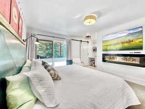 Un dormitorio moderno con una cama grande, chimenea, televisor y vista a un paisaje por la ventana.