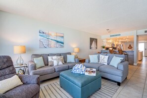 Ein Wohnzimmer mit einer grauen Sofagarnitur, einem blauen Hocker und einem Bild von einer Strandansicht.