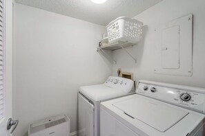 A laundry room with a front-loading washer and dryer, a white laundry basket on a shelf, and a vent on the floor. - Anna Maria Island Club Unit 38 (Bradenton Beach)