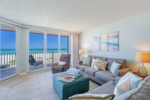 A spacious living room with a balcony overlooking the ocean, a grey sofa set, and a large window. - Anna Maria Island Club Unit 38 (Bradenton Beach)
