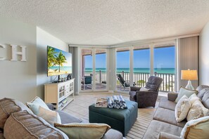 Ein großzügiges Wohnzimmer mit einem großen Fernseher, bequemen Sitzgelegenheiten, Meerblick und einem Balkon mit Gartenmöbeln.