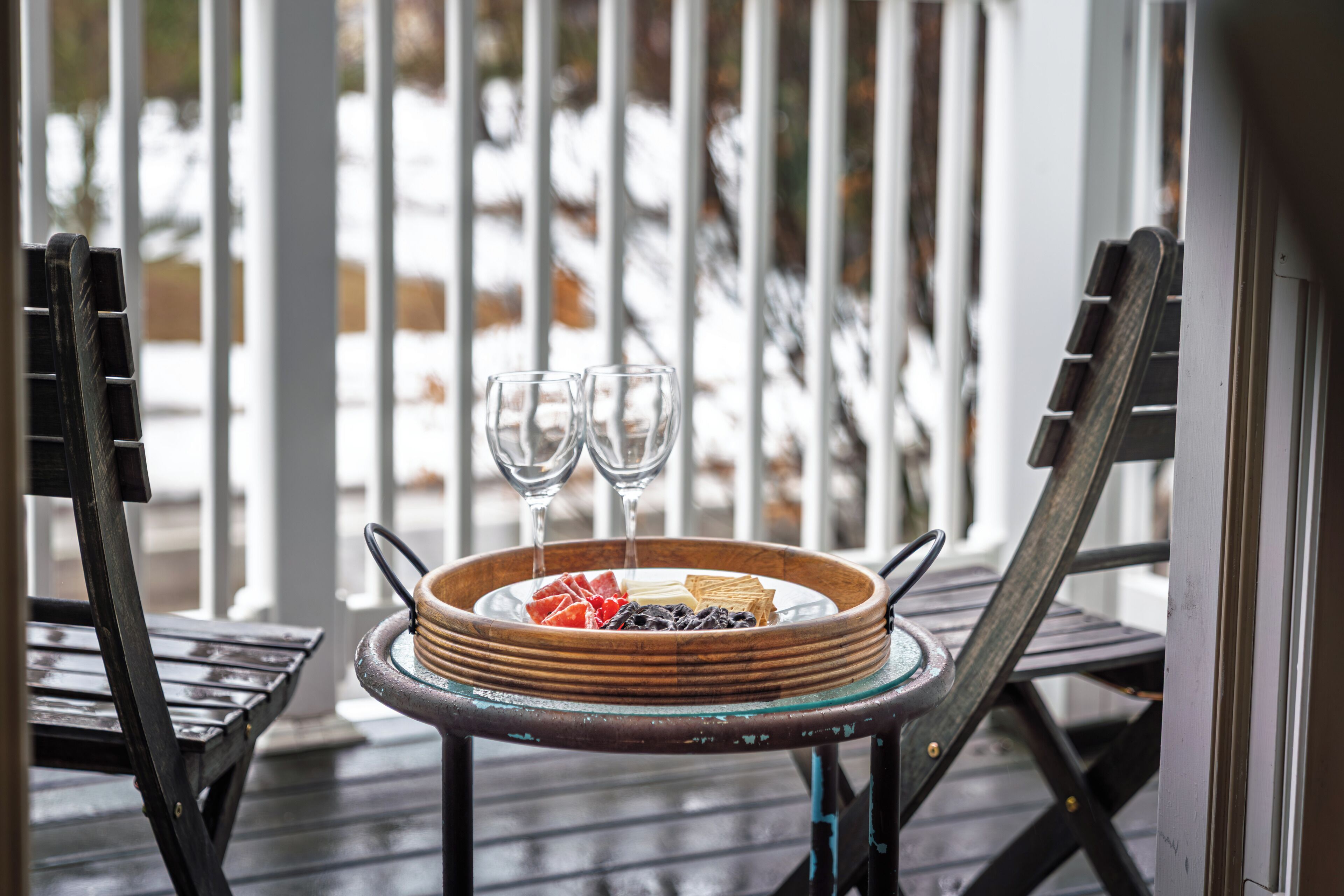 A wooden table with a glass bowl containing food and two glasses of water, set on a porch with a railing.