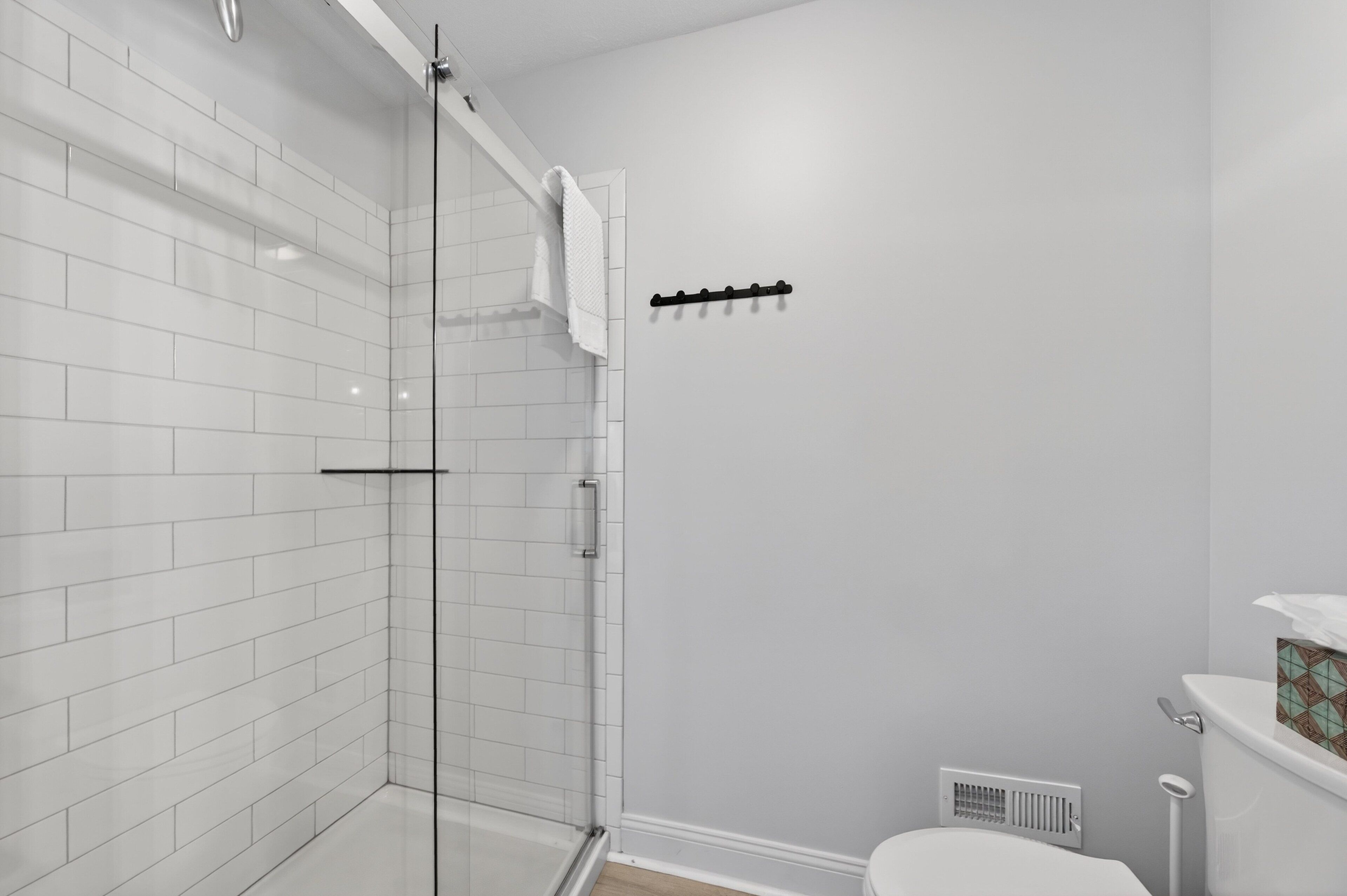 Une salle de bain avec des murs recouverts de carreaux blancs, une douche en verre, des toilettes et un porte-serviettes.