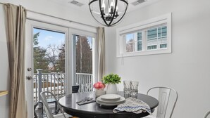 Uma área de refeições com uma mesa redonda, cadeiras, um candelabro, uma janela com persianas e vista para árvores.