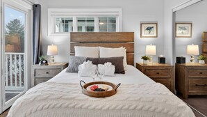 Cama bem arrumada com uma bandeja de frutas, duas luminárias laterais e vista para o exterior através de uma grande janela.