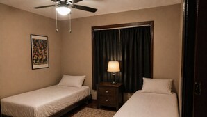 A bedroom with two single beds, a ceiling fan, a nightstand with a lamp, a framed picture on the wall, and a window with curtains.